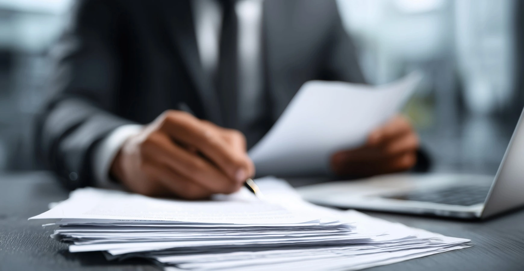 Text: Close-up of a business professional in a suit reviewing and signing official visa paperwork on a desk next to a laptop.